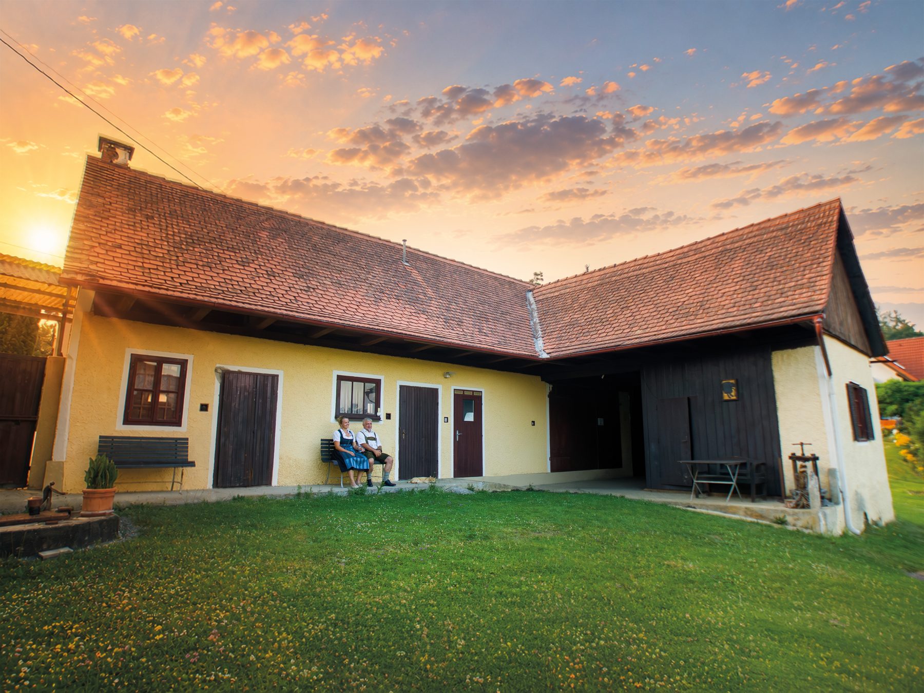 Maria und Christoph sitzen im Abendlicht vor dem gelben Vierkanthof